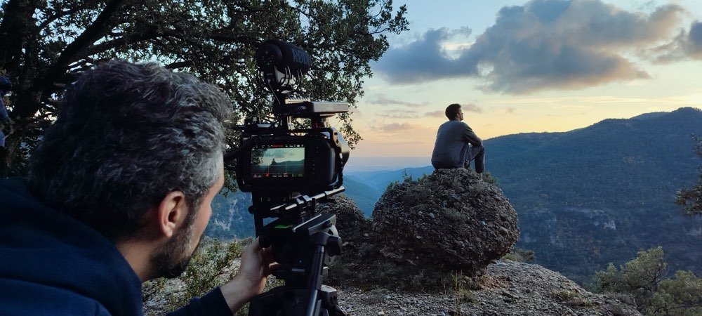Un cámara del equipo filma a un joven sentado sobre una roca frente a las montañas, al atardecer.